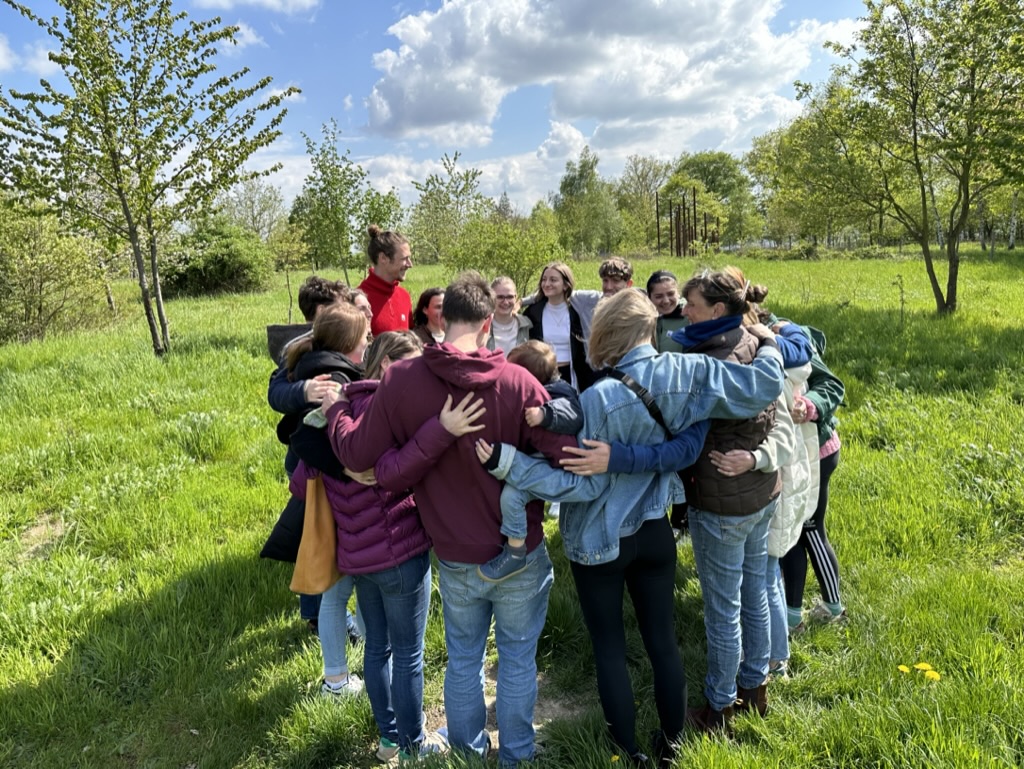 Menschen stehen in einer grünen Wiese eng im Kreis und legen die Arme umeinander.